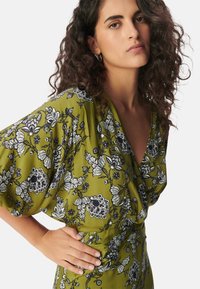 Vestido floral verde con patrones en blanco y negro, mangas cortas tipo murciélago y un diseño cruzado que acentúa la cintura. Textura de tela suave.