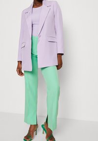 Blazer ajusté lavande avec revers, pantalon vert menthe avec fentes latérales, et escarpins verts à lanières. Tissu lisse et coupe structurée.