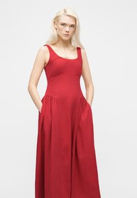 Νεαρή γυναίκα με μακριά ξανθά μαλλιά, φορώντας αμάνικο κόκκινο μακρύ φόρεμα με τσέπες, στέκεται μπροστά σε απλό φωτεινό φόντο.