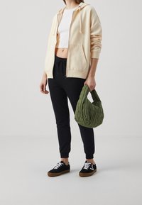 Mujer con sudadera beige de cremallera, top corto blanco, pantalones jogger negros, zapatillas negras, sosteniendo un pequeño bolso acolchado verde oliva.