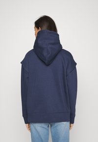 Felpa con cappuccio blu navy, con maniche larghe, texture morbida e tasca a marsupio sul davanti. La vista posteriore mostra il cappuccio e la vestibilità rilassata.