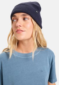 Tengerészkék kötött sapka bordázott mintával és kis logóval; párosítva egy kék pamut pólóval, amelynek kerek nyaka és rövid ujjai vannak.