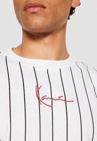 White t-shirt with black vertical stripes, featuring red embroidered text. Crew neck design, cotton material, close-up view.