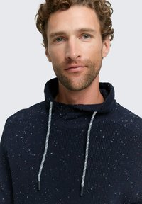 Marineblaue gestrickte Kapuzenjacke mit einem strukturierten Muster, das mehrfarbige Sprenkel enthält und einen hohen Kragen hat. Sie verfügt über silberne Kordeln mit schwarzen Spitzen.
