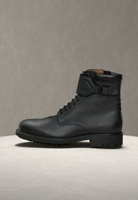 Bottine en cuir noir à lacets à l'avant, détail de bride à boucle, col rembourré et semelle robuste sur une surface claire avec un fond gris.