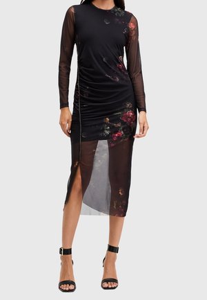 Vestido negro de malla con mangas largas, estampado floral, detalle fruncido, abertura alta lateral, combinado con tacones negros con pulsera en el tobillo.