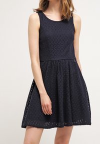 Robe sans manches bleu marine avec un encolure ronde, en tissu dentelle texturée, corsage ajusté et jupe évasée s'étendant jusqu'au mi-cuisse.