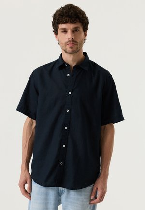 Mann mit lockigem Haar, der ein schwarzes kurzärmeliges Hemd mit Knöpfen und hellblaue Jeans trägt, steht vor einem schlichten hellen Hintergrund.