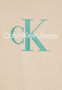 Bežna pamučna tkanina s grafičkim dizajnom koji uključuje "Calvin Klein Jeans" u bijeloj i tirkiznoj boji. Ističe se hrabrom tipografijom i kontrastom boja.