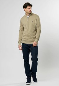 Beige hettejakke med glidelås, høy krage, ribbede mansjetter og en liten logo på brystet, sammen med mørkeblå jeans og marineblå joggesko.