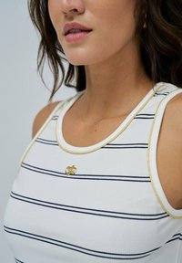 Camiseta sin mangas blanca con rayas en azul marino, borde dorado en el cuello y los brazos, con un pequeño logo dorado en el pecho. Textura acanalada.
