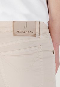 Pantaloni di cotone beige con superficie testurizzata, dotati di una etichetta in pelle con la scritta "JECKERSON" stampata sul retro della vita e cuciture dettagliate.