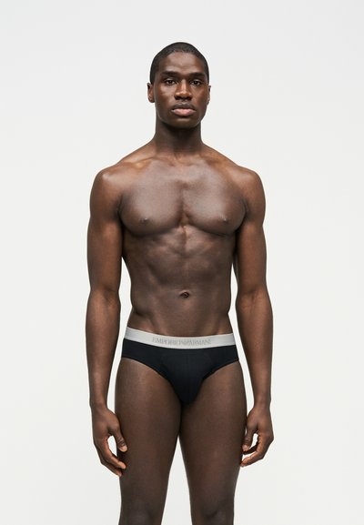 Μυώδης άνδρας στέκεται ακίνητος φορώντας μαύρα εσώρουχα Emporio Armani με λευκή ζώνη μέσης, μπροστά σε απλό ανοιχτό φόντο.