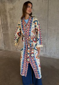 Γυναίκα που φοράει ένα πολύχρωμο παλτό με γεωμετρικό μοτίβο και ζώνη, μπλε τζιν και κρατά μια μπεζ σουέντ τσάντα μπροστά σε ένα τσιμεντένιο τοίχο.