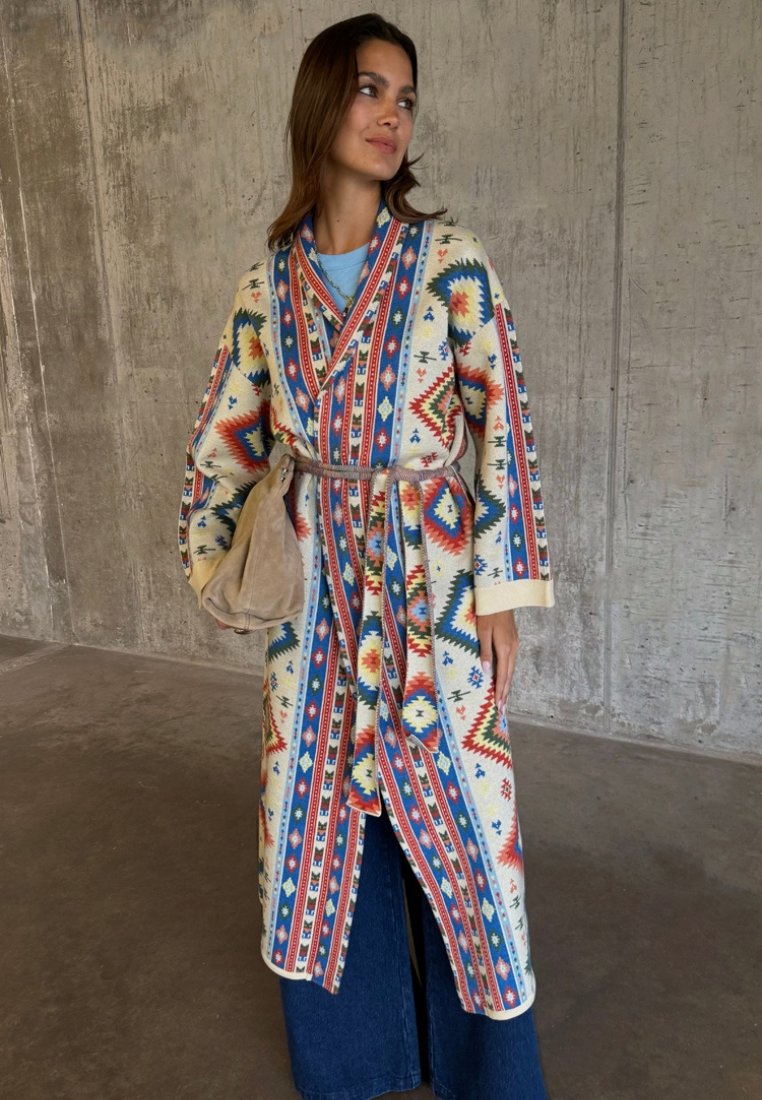 Γυναίκα που φοράει ένα πολύχρωμο παλτό με γεωμετρικό μοτίβο και ζώνη, μπλε τζιν και κρατά μια μπεζ σουέντ τσάντα μπροστά σε ένα τσιμεντένιο τοίχο.