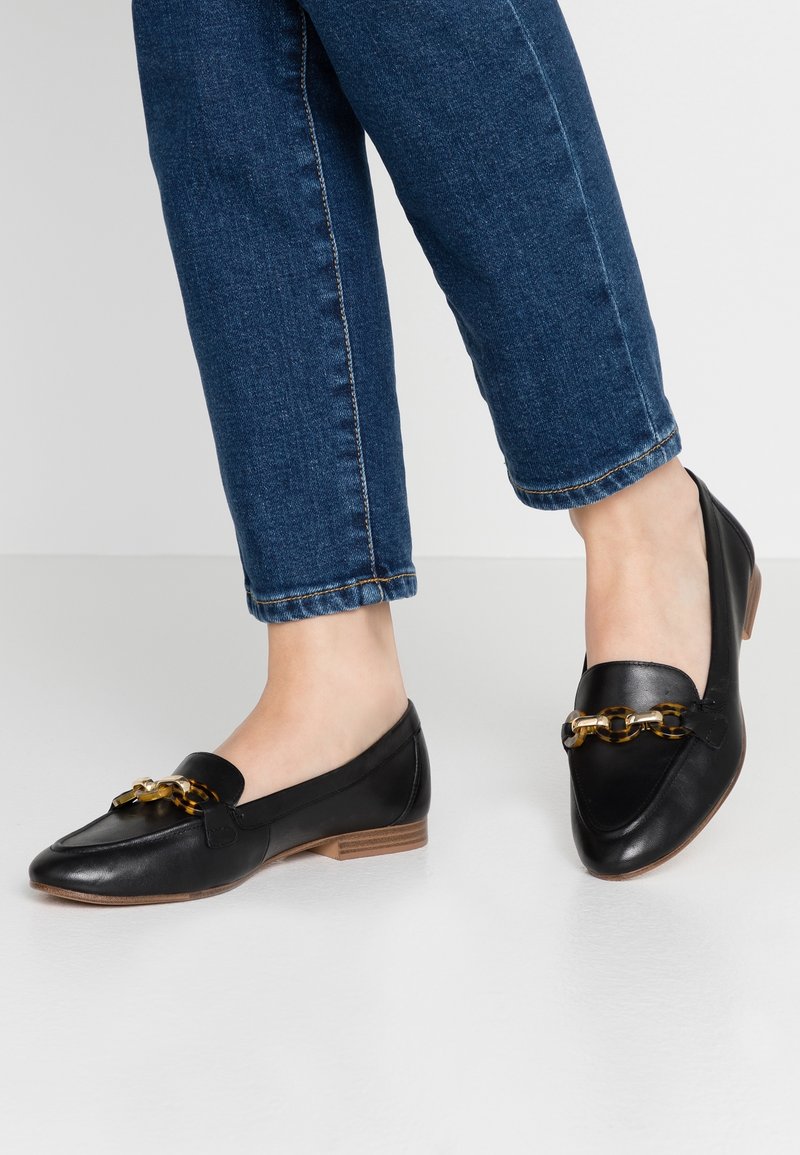 Mocassins en cuir noir avec détail de chaîne en écaille portés avec un jean en denim bleu sur un sol clair.