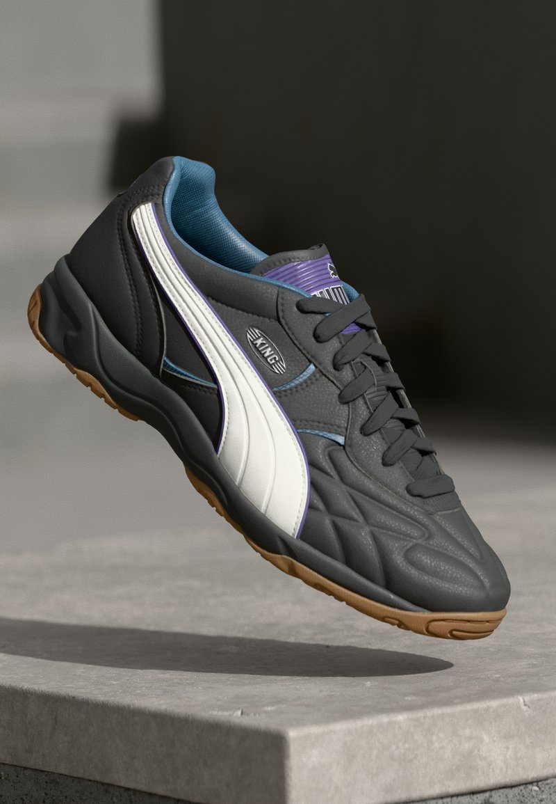 Szürke sportcipő textúrált felsőrésszel, fehér oldalcsíkkal, lila részletekkel és kék belsővel. Rágógumin talp.