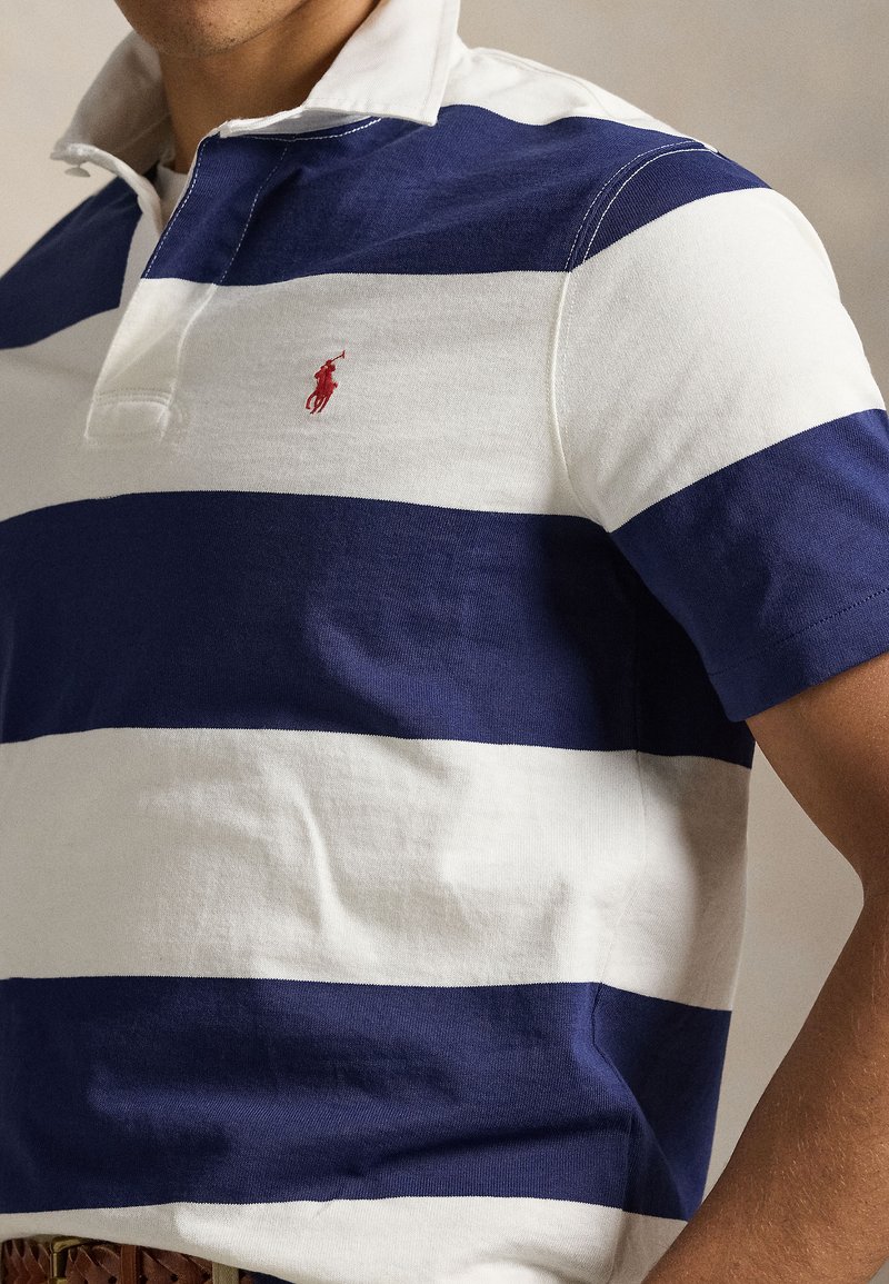 Polo rayé alternant bleu marine et blanc, manches courtes, col blanc, avec un petit logo rouge sur la poitrine.