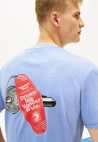 Jasnoniebieska bawełniana koszulka z grafiką czerwoną metkę z napisem "Marz o wielkim, wędruj szeroko" na plecach, okrągły dekolt i krótkie rękawy.