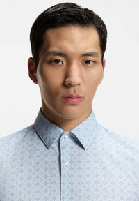 Jonge man met kort zwart haar, gekleed in een lichtblauw overhemd met patroon en knopen, kijkt recht in de camera tegen een effen achtergrond.