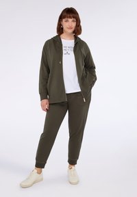 Olivově zelená mikina s zipem, spárovaná s bílým tričkem s potiskem a odpovídajícími olivovými tepláky. Bílé tenisky dokončují outfit.