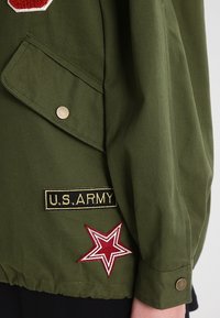 Oliwkowa kurtka wojskowa w stylu z złotymi akcentami, z czarną naszywką "U.S. ARMY" i haftem czerwonej gwiazdy na rękawie.