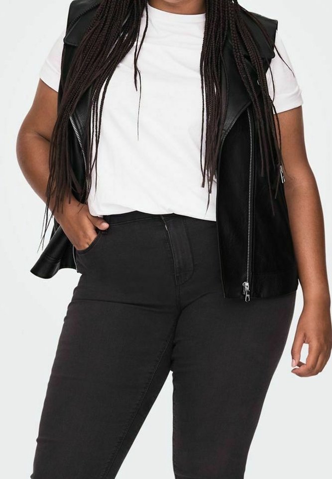 Černá vesta z umělé kůže s rameními vycpávkami, nošená přes bílý tričko, v kombinaci s vysokými černými džíny. Viditelné dlouhé zapletené vlasy.
