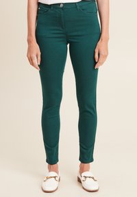 Jeans skinny verde in denim con una texture liscia, dotati di tasca frontale, chiusura con bottone e passanti per cintura, abbinati a scarpe bianche senza lacci.