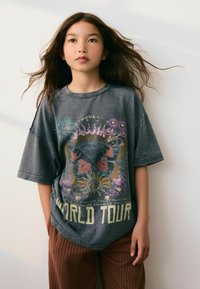 Szürke, oversized póló színes pillangó mintával és "WORLD TOUR" felirattal. Bőrszínű bordás nadrággal párosítva, semleges háttér előtt.