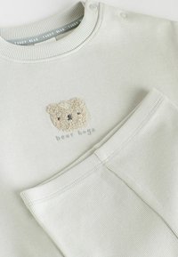 Ljust grå bomullströja och matchande byxor har en texturerad teddy-björn patch och texten "björnkramar" i mjuka färger. Ribbad halsringning och muddar.