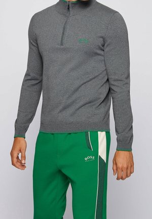 Grijze zip-up trui met een hoge kraag, geribbelde mouwen en een groen logo op de borst, gecombineerd met groene sweatpants met een witte streep.