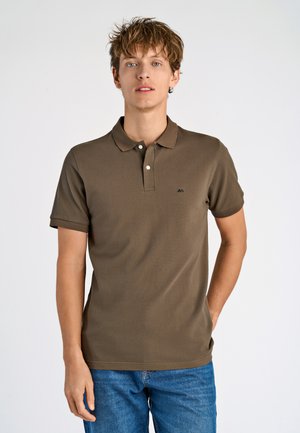 Camisa polo de algodón marrón con un corte ajustado, mangas cortas, tapeta de dos botones y un pequeño logo negro en el lado izquierdo del pecho. Usada con jeans azules.
