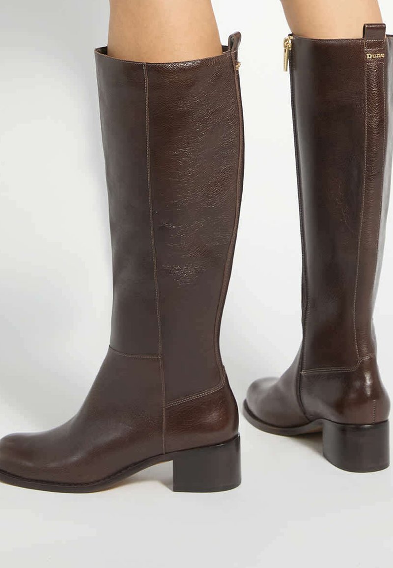 Bottes en cuir marron montant jusqu'aux genoux avec des petits talons blocs portées par une personne debout sur un fond clair.