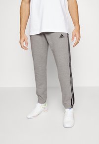 Grå sweatpants med svarta sidostråkar och Adidas-logotyp. Tillverkade av ett mjukt material, med en tapered design och elastisk midja.