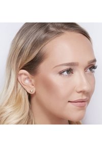 Gouden hartvormige studs op een model, met een gladde metallic afwerking en licht afgeronde randen.
