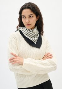 Kvinde med mørkt krøllet hår iført en lys cremet kabelstrikket sweater og et sort-hvidt stribet tørklæde, med armene korslagte, mens hun kigger til siden.