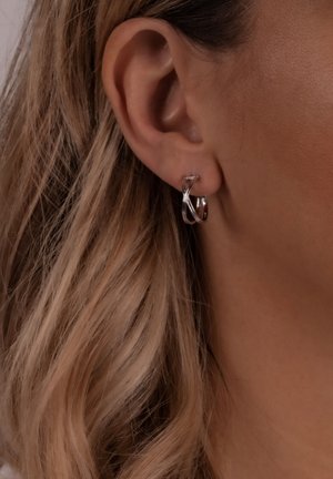 Pendientes de aro de plata con un diseño entrelazado, que presentan un acabado pulido. Llevados en la oreja de un modelo, combinados con un cabello rubio ondulado.
