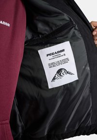 Innenansicht einer schwarzen Jacke mit weißem Pegador-Label mit Berglogo und Text über Outdoor-Bekleidung für abenteuerlichen Gebrauch.