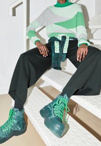 Pull en maille à motifs verts et gris avec un col rond, associé à un pantalon noir à jambes larges et des bottines vertes à lacets, posé sur un escalier.