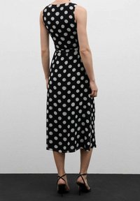 Robe noire sans manches avec des pois blancs, longueur mi-mollet, taille cintrée et tissu fluide, associée à des escarpins noirs à lanières.