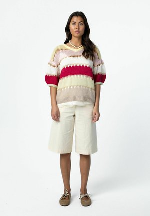 Kvinde stående iført en stribet striktrøje med røde, hvide, beige og lyserøde farver, cremekorte shorts og brune snørestøvler.