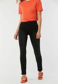 Jean noir taille haute associés à un t-shirt orange vif et des sandales à talons rouges avec un design en maille. Tissu lisse et coupe ajustée.