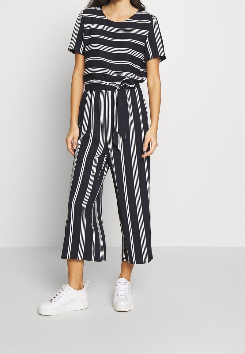 Vrouw in een zwart-wit gestreepte jumpsuit met een strikceintuur en witte sneakers, staand tegen een effen lichtgrijze achtergrond.