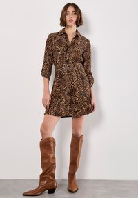 Robe chemise à imprimé léopard dans des tons bruns avec un col, une fermeture à boutons à l'avant et une taille ceinturée. Associée à des bottes en cuir marron jusqu'aux genoux.