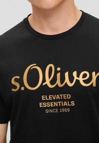 Schwarzes Baumwoll-T-Shirt mit großem goldfarbenem Druck von "s.Oliver" und "ELEVATED ESSENTIALS" mit "SINCE 1969" darunter. Runder Ausschnitt und kurze Ärmel.