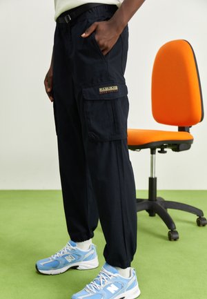 Osoba ubrana w czarne spodnie cargo i niebieskie sneakersy New Balance stoi obok pomarańczowego krzesła biurowego na zielonej podłodze.