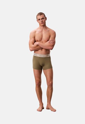 Hombre musculoso de pie descalzo con los brazos cruzados, vistiendo bóxers Calvin Klein de color verde oliva sobre un fondo blanco liso.