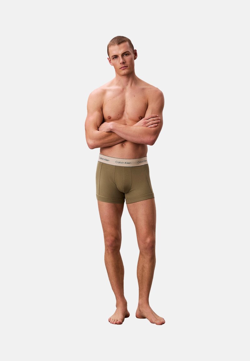 Homme musclé debout pieds nus, les bras croisés, portant un boxer Calvin Klein vert olive sur un fond blanc uni.