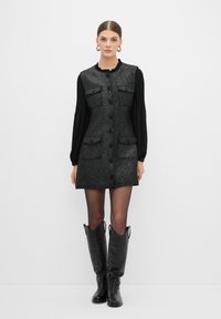 Vestido sin mangas de tweed negro con dos bolsillos frontales, cinco botones y un cuello con volantes, combinado con botas de cuero negras hasta la rodilla.