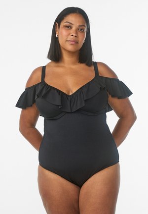 Selvsikker kvinde iført en sort off-shoulder badedragt med flæsedetaljer, stående med hænderne bag ryggen.